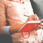 Close-up of a woman writing in a red journal on a sofa, wearing a stylish ring.